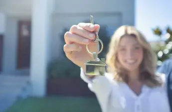 woman-smiling-and-holding-house-key-towards-camera-while-standing-in-front-of-house