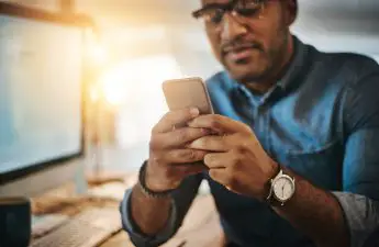 man-sitting-at-desk-with-computer-while-using-smartphone-app