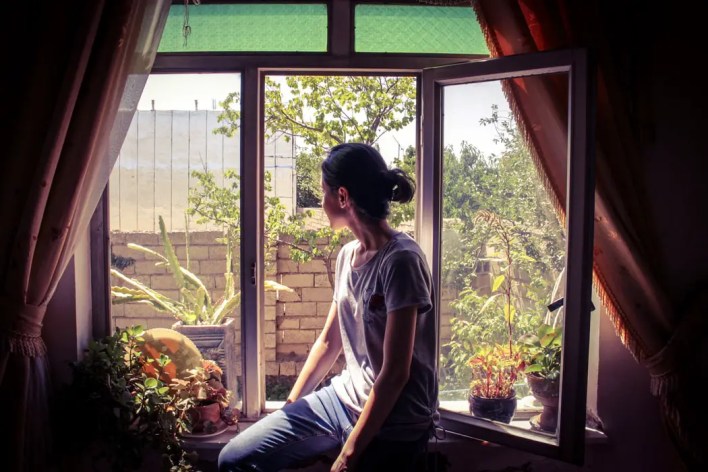 young-woman-sitting-on-ground-floor-windowsill-inside-rented-property-and-looking-outside-at-trees